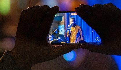 Hands holding an iphone, recording a performer onstage with a microphone.