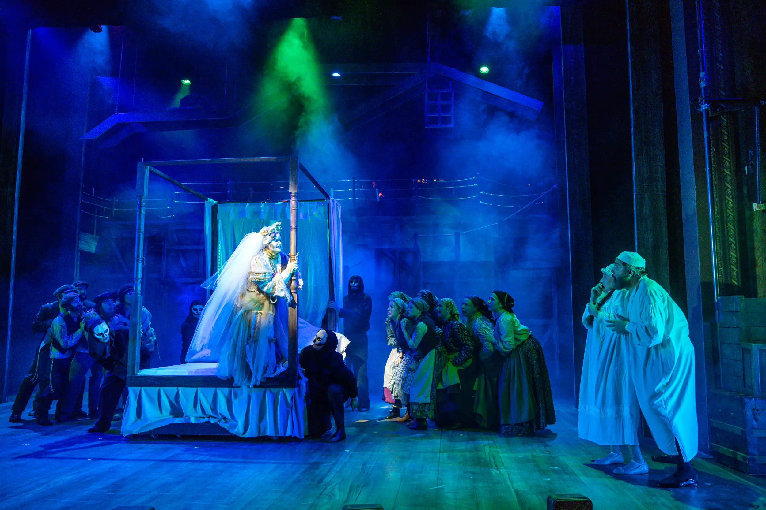Cast on stage for a performance. Woman on bed with crowd around her.