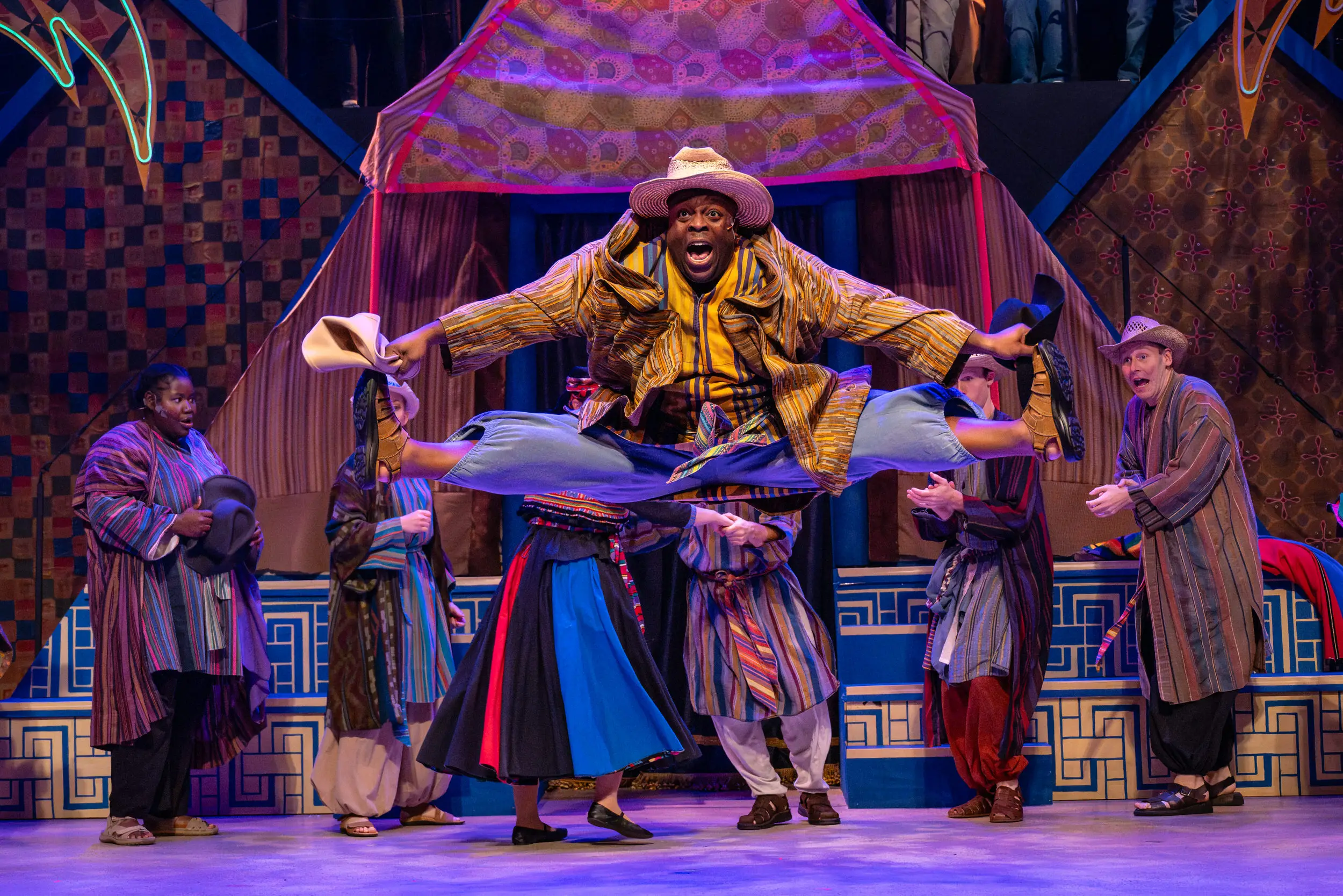 Man jumping and doing splits mid air with cast behind him on stage for performance of Joseph and the Technicolor Dreamcoat