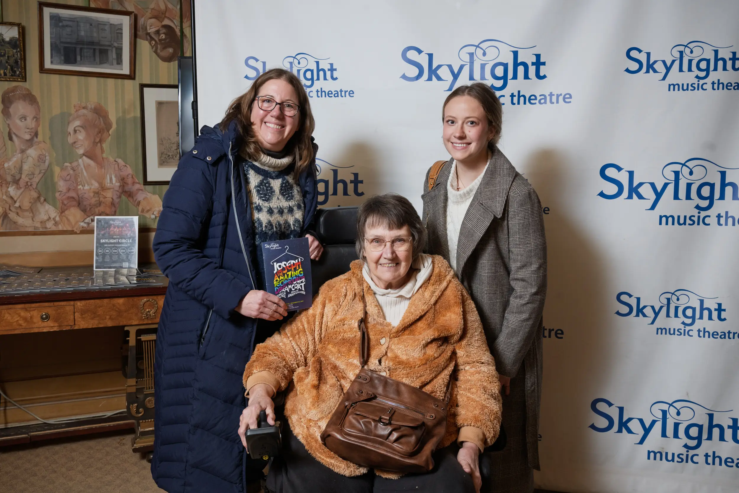 Three women and a playbill in front of the Skylight backdrop